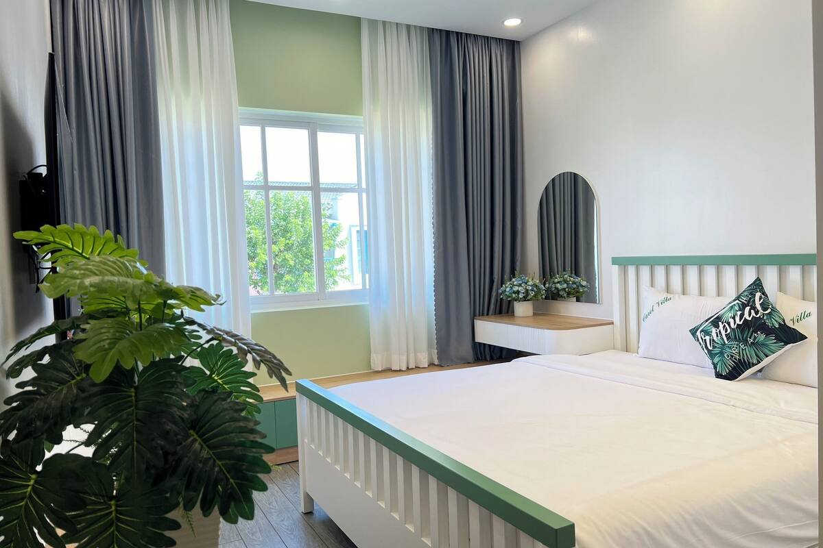 A bedroom with a bed, a bedside table with a mirror, a potted plant, and a window with curtains.