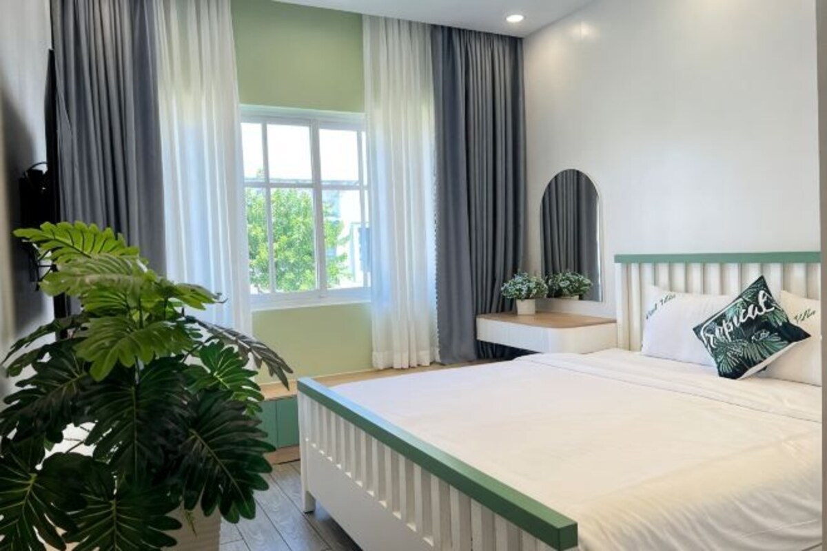 Ein Schlafzimmer mit Bett, Nachttisch, Spiegel, Fenster mit Vorhängen und einer Topfpflanze.