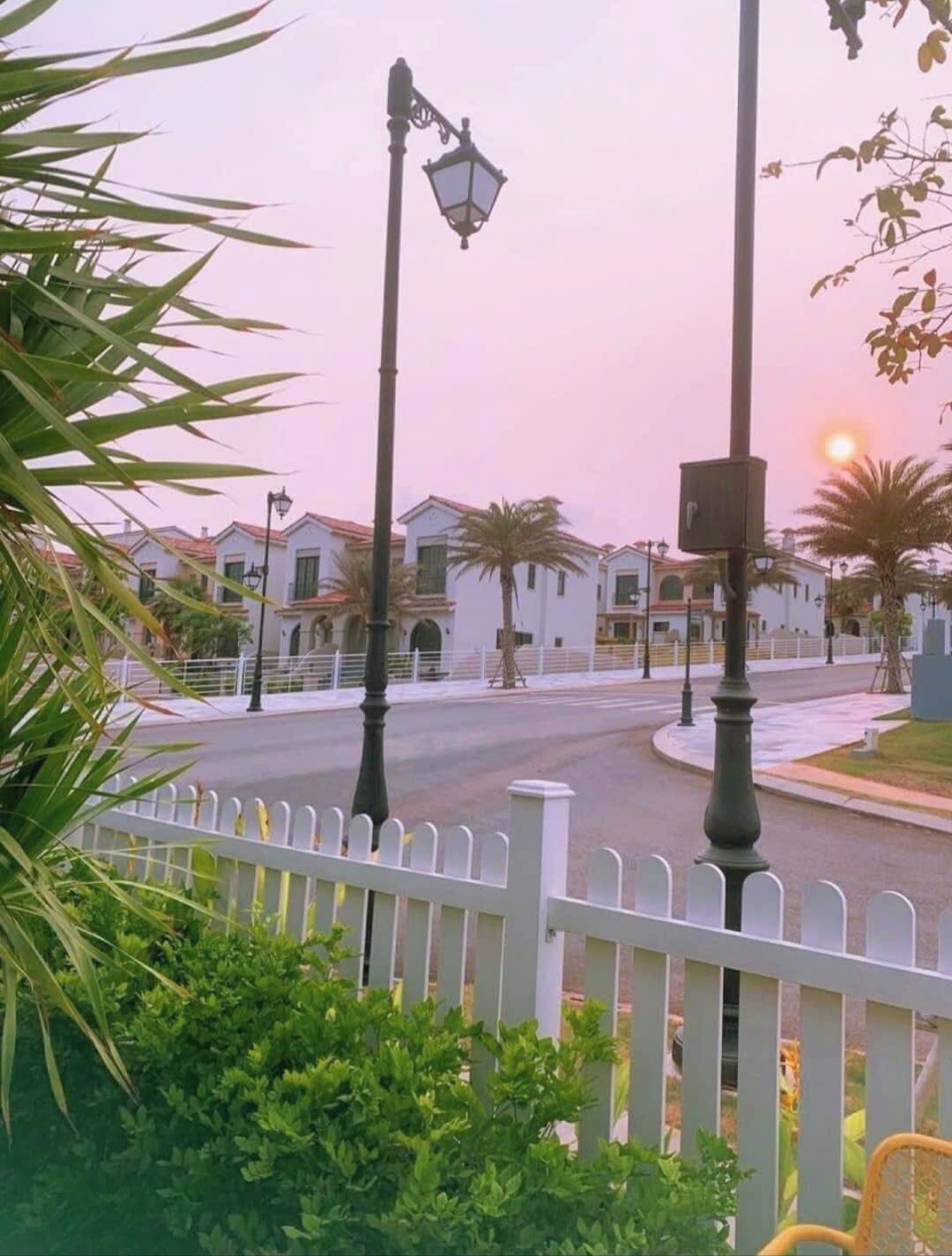 A street view with a white picket fence, street lamps, and a row of white buildings with red roofs.