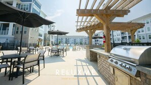An outdoor patio with tables and chairs, a barbecue grill, and a pool in the background. - Reluxme|Chic  2BR w/ Balcony, Gym & Pool Near Mall (Charlotte)