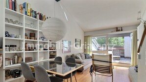Un salon moderne avec une table à manger en verre, des chaises grises, un canapé et une bibliothèque remplie de livres et d’objets décoratifs.