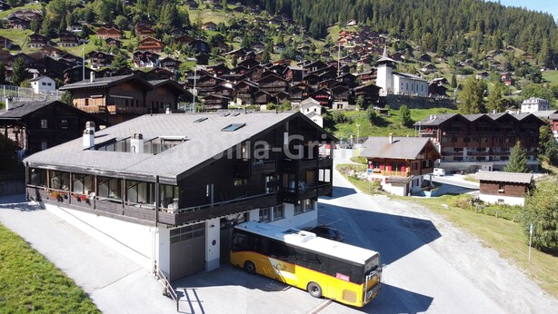 Un bâtiment moderne, avec un autobus garé devant, entouré de maisons traditionnelles en bois sur une colline.