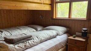 Una cama litera con cabecera de madera, una mesita de noche con lámpara y una ventana con vista a árboles.