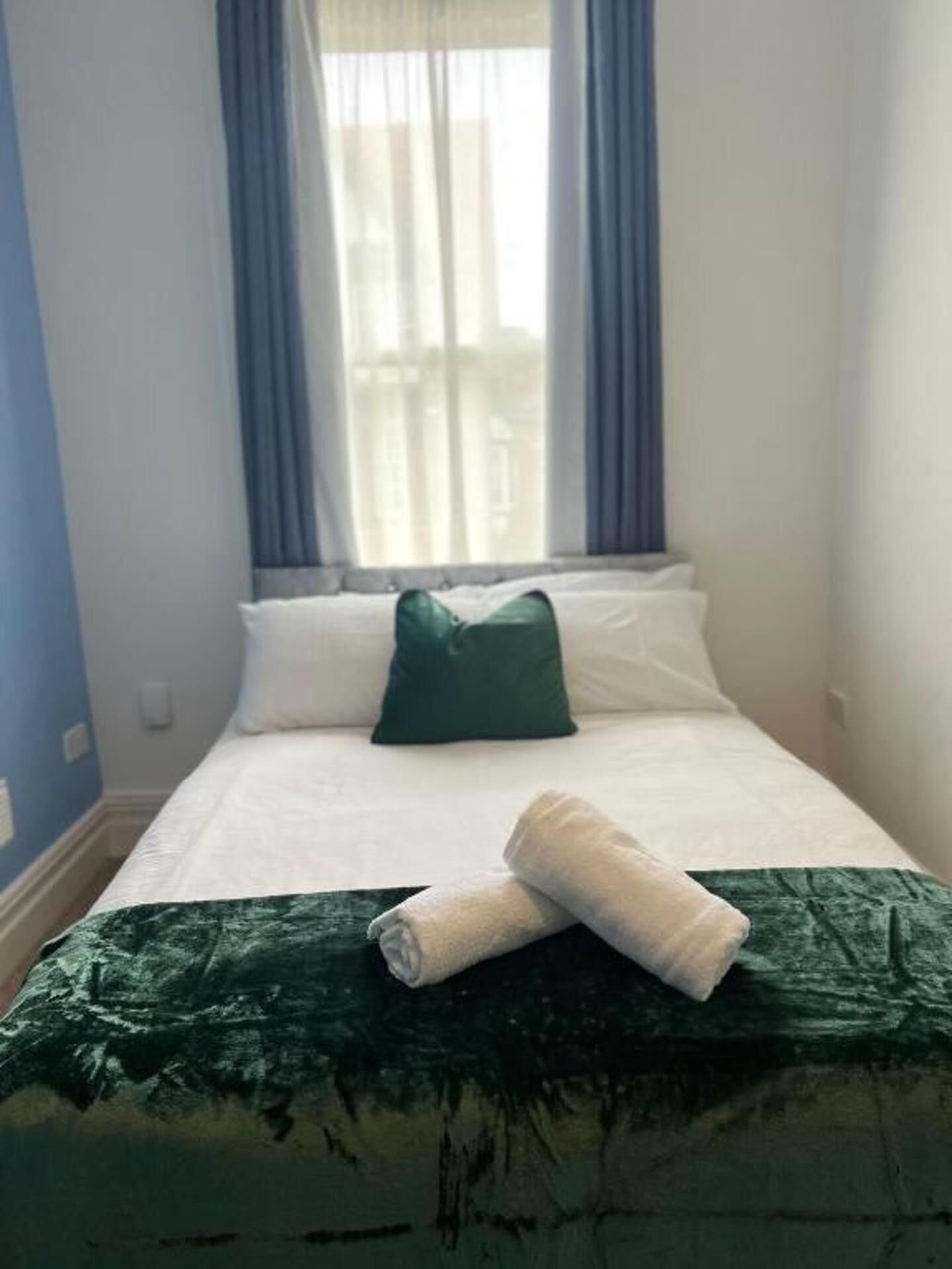 Una cama bien hecha con una colcha verde y una toalla enrollada, un almohadón blanco y un almohadón verde sobre una pared azul con una ventana y cortinas.