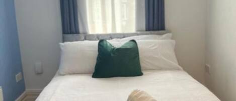 Una cama bien hecha con una colcha verde y una toalla enrollada, un almohadón blanco y un almohadón verde sobre una pared azul con una ventana y cortinas.