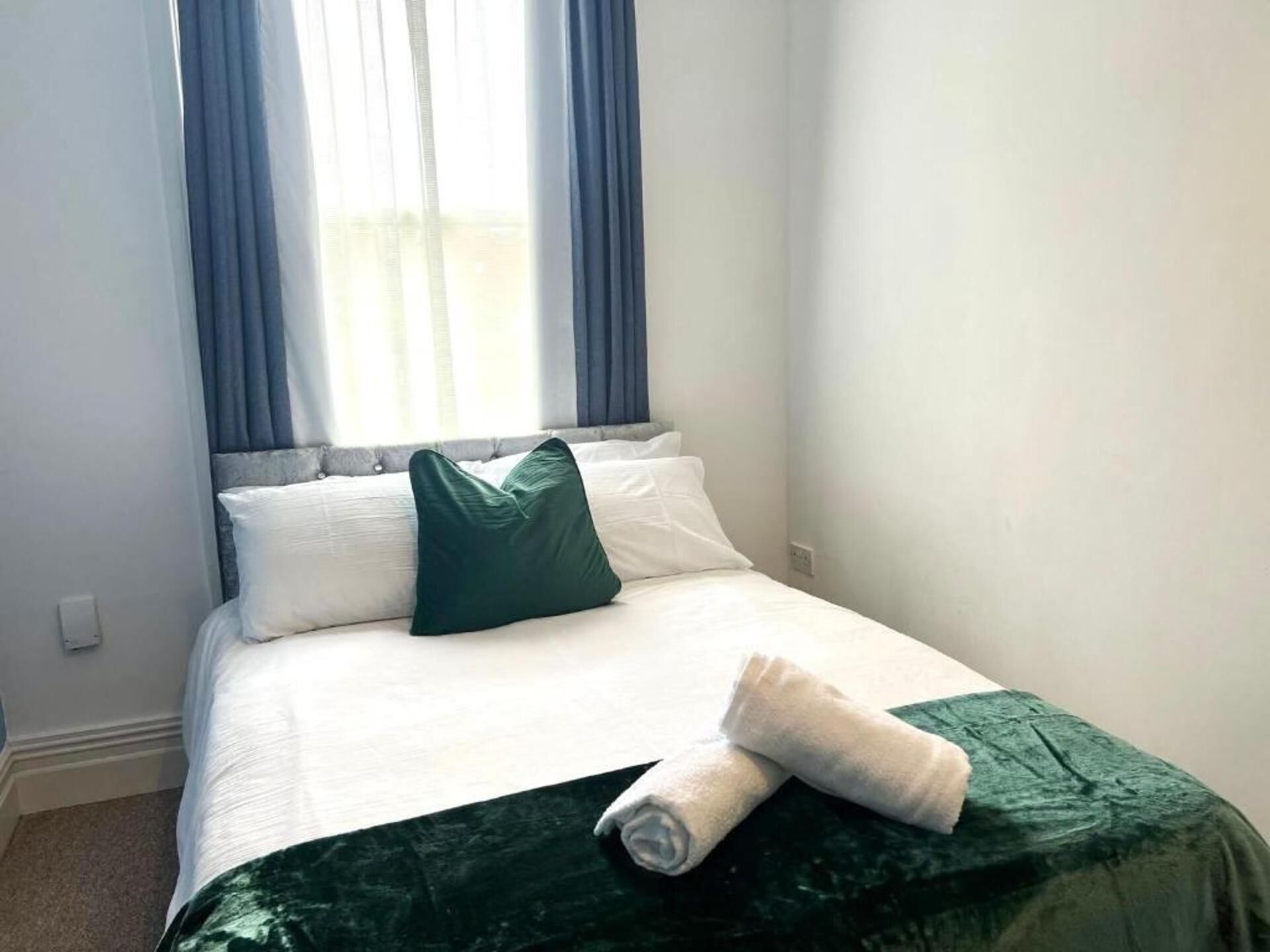 Een netjes opgemaakt bed met witte lakens, een groen kussen, een opgerolde witte handdoek en een donkergroene plaid.