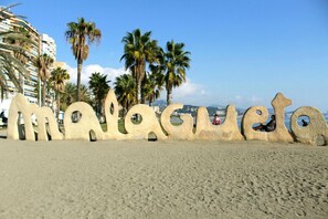 Une plage où de grandes lettres en pierre, formant des motifs abstraits, épellent le mot « Malagueta », avec des palmiers en arrière-plan.