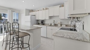 Een moderne keuken met witte kasten, granieten aanrechtbladen en een ingebouwde kookplaat. Er staan barkrukken en er is een raam met uitzicht op een balkon.