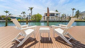 Een houten terras met twee witte loungestoelen en een kleine tafel, met uitzicht op een waterpartij en een rij huizen.