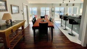 A dining area with a wooden table and chairs, a bench, a lamp, and a view of the sea through large windows.
