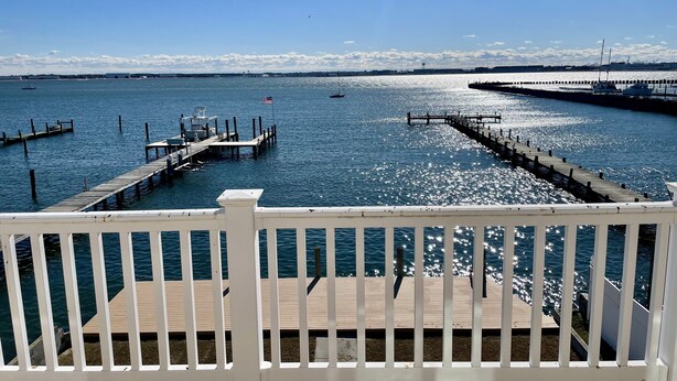 A waterfront view from a deck with a wooden railing, a dock extending into the water, and a clear sky.