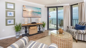 A living room with a large painting of a beach, a ceiling fan, and a view of the ocean through a sliding glass door. - Phoenix Iii 3084 2 Bedroom Condo (Orange Beach)