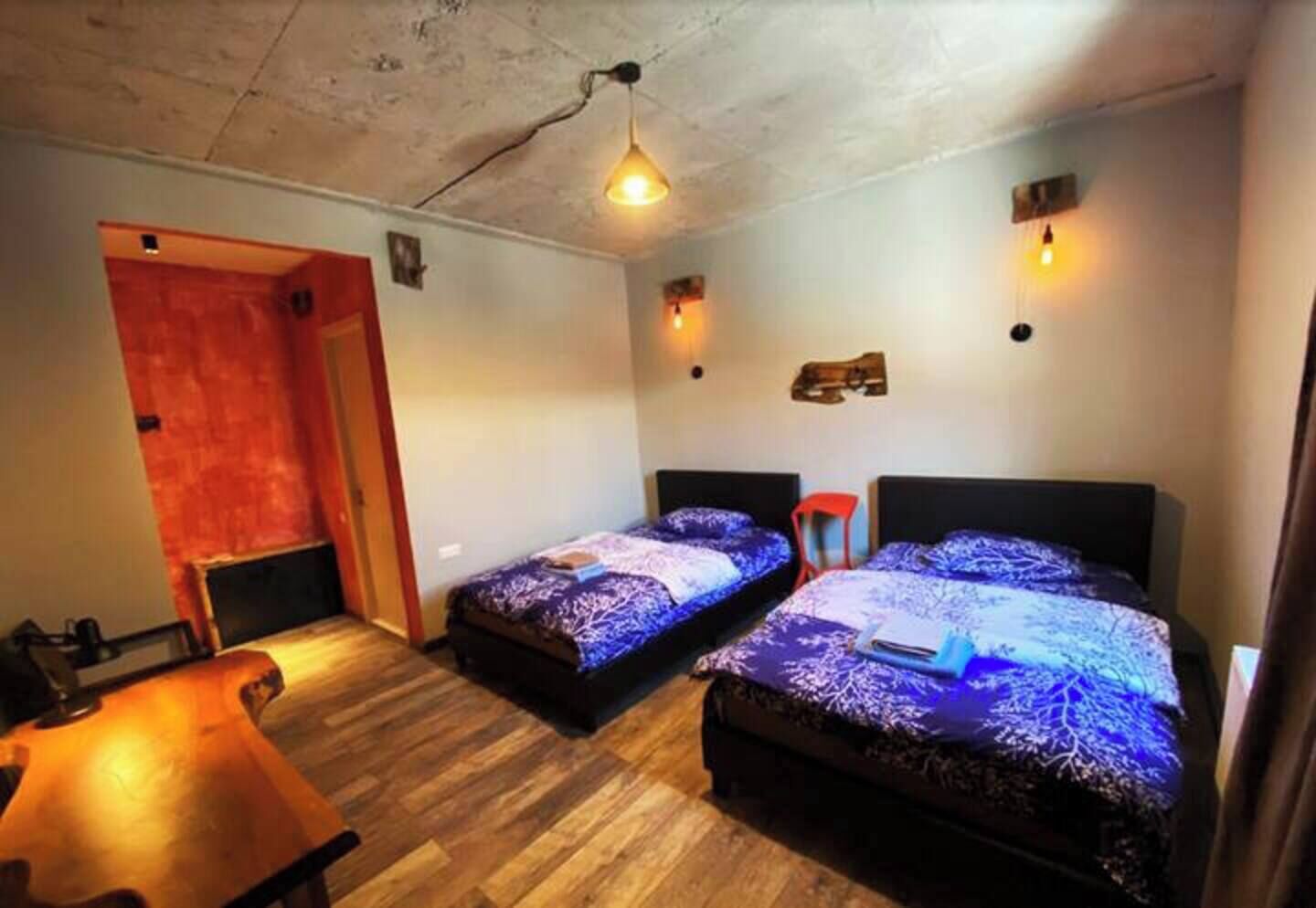 A small, cozy room with two beds, a wooden table, and a red door.