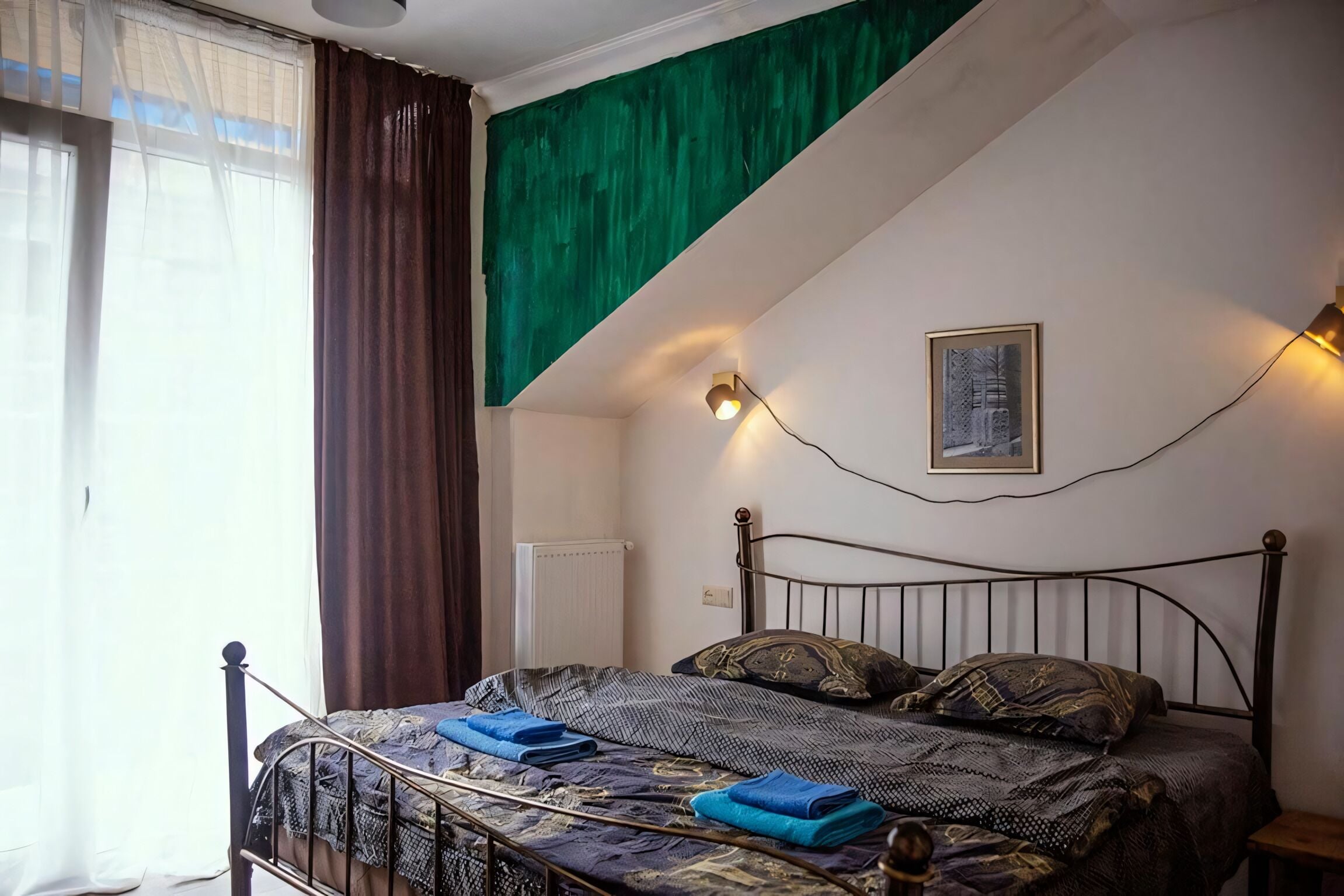 Une chambre avec un lit, un radiateur, une fenêtre avec des rideaux, un mur d’accent vert et un luminaire fixé au mur.