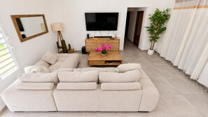 Una sala de estar con un sofá beige grande, una mesa de centro de madera con flores rosadas, un televisor de pantalla plana y una decoración de cactus.