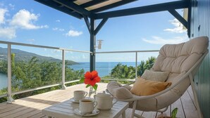 Ein Balkon mit einem weißen Sitzmöbel, einem kleinen Tisch mit einer Blume und Blick auf das Meer und die Berge.