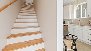 Una escalera con escalones de madera y paredes blancas que conduce a una cocina con armarios blancos y un comedor con una mesa y sillas.