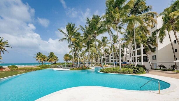 Un espace piscine aménagé dans un complexe hôtelier, avec des palmiers, une vue sur la plage et un ciel dégagé.