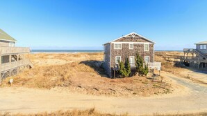 A two-story house with a wooden deck, situated on a sandy beach with dunes in the background. - Ocean Views, Gorgeous! Dogs Welcome, Hot Tub, SN9805 (Nags Head)