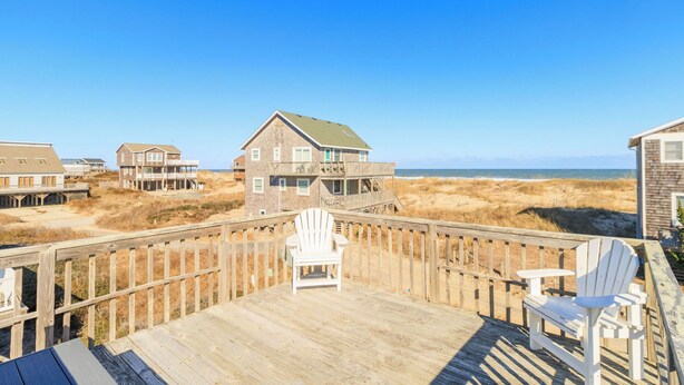 A wooden deck with two white Adirondack chairs, overlooking a beach with multiple houses and dunes. - Ocean Views, Gorgeous! Dogs Welcome, Hot Tub, SN9805 (Nags Head)