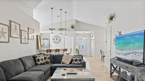 Ein modernes Wohnzimmer mit Ecksofa, Couchtisch und Fernseher auf einem Holzboden. An der Wand hängen gerahmte Bilder, eine Uhr und von der Decke hängt eine Pflanze.