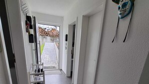 A hallway with a tiled floor, a white wall with a decorative bird sculpture, and a door leading to an outdoor area with a fire hydrant and stacked firewood. - Holiday Apartment Moises y Ruth with beautiful garden (Murg)