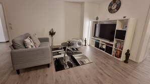 A modern living room with a grey sofa, a black coffee table, a round side table, a black and white rug, a flat-screen TV on a white stand, and a bookshelf with books and decorative items. - Holiday Apartment Moises y Ruth with beautiful garden (Murg)