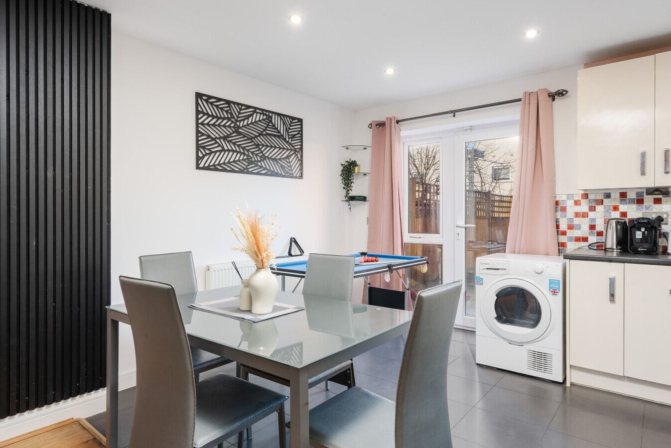 A modern kitchen with a washing machine, a dining area with a glass table, and a pool table.