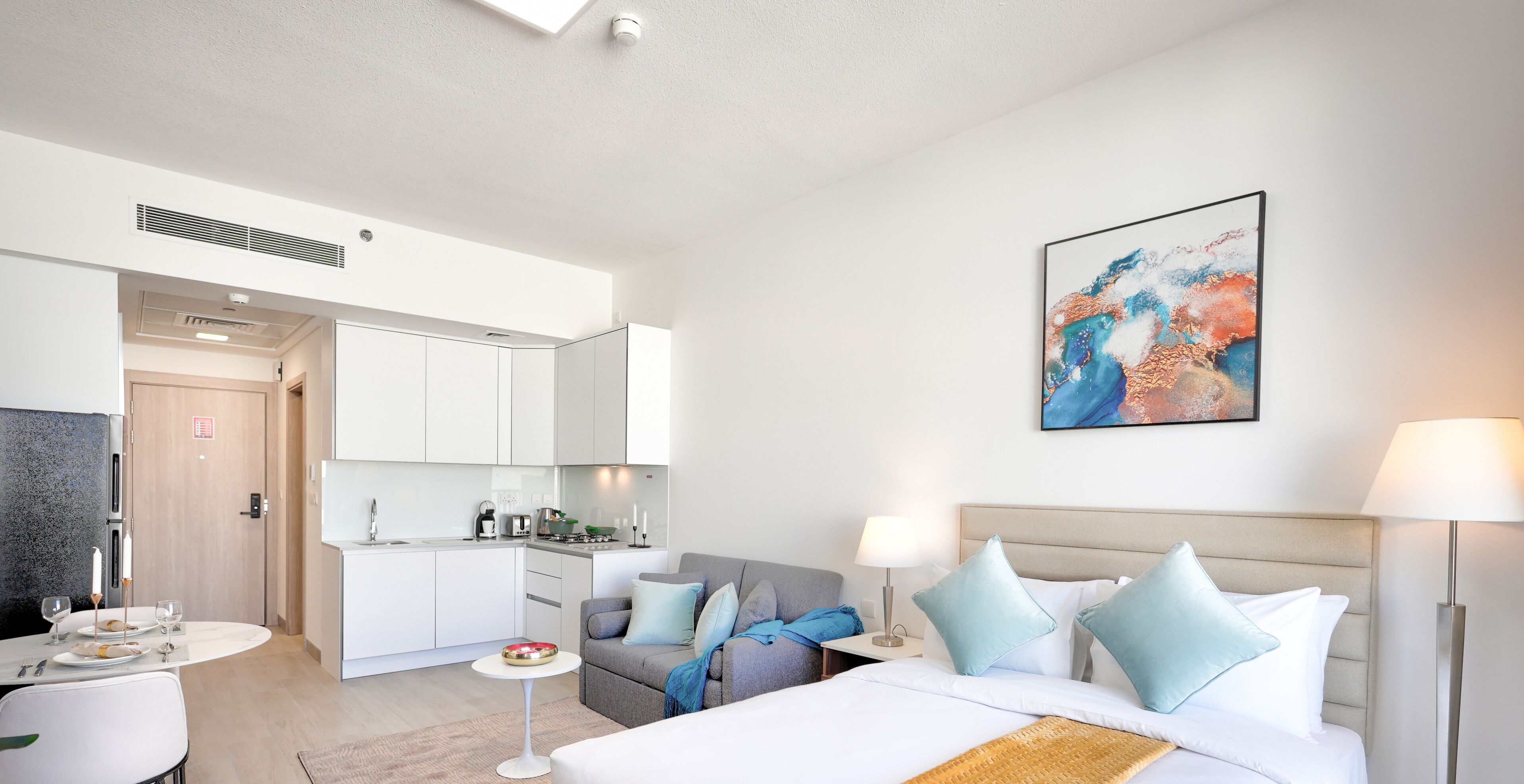 A modern hotel room with a bed, bedside tables, a sofa, a dining area, and a kitchen.