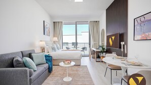 Ein modernes Hotelzimmer mit grauem Sofa, einem runden Couchtisch, einem Bett mit weißem Kopfteil, einem Balkon mit Aussicht und einer Essecke für zwei Personen.
