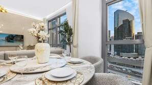 A modern dining area with a round table set for two, a vase with white flowers, and a city view through large windows. - Walk to Dubai Mall | 2BR Apartment with Balcony and Pool (Downtown)