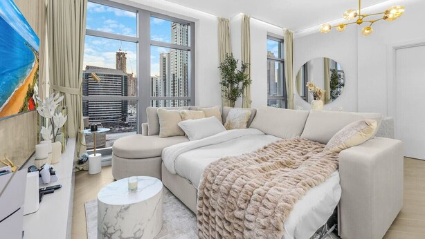 A modern bedroom with a large bed, a seating area, and a view of the cityscape. - Walk to Dubai Mall | 2BR Apartment with Balcony and Pool (Downtown)