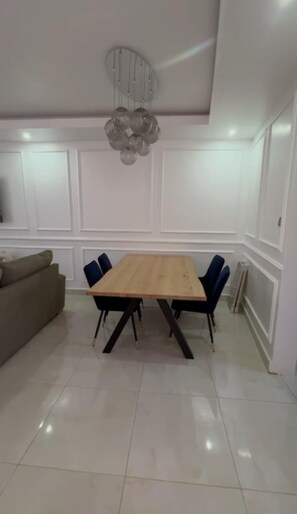 Un espace repas avec une table en bois, des chaises bleues, un lustre et des murs recouverts de panneaux blancs.