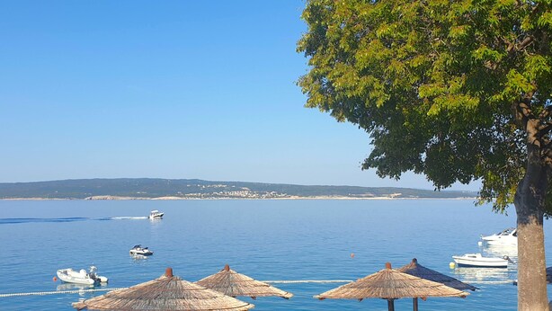 Une vue sur la côte, avec un ciel dégagé, une mer calme et plusieurs bateaux. On y voit des parasols en chaume et un arbre qui offre de l’ombre.