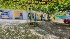 Un chemin pavé mène à un bâtiment à la façade blanche et à la porte jaune. Des voitures sont garées sur le côté, et une voiture rouge se trouve à l’arrière-plan.