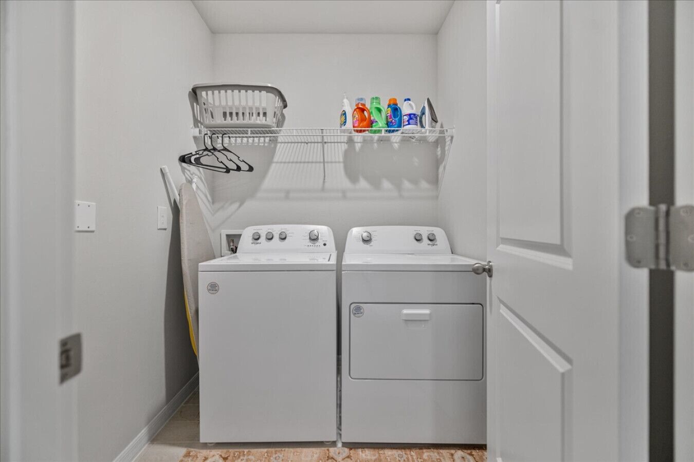A laundry room with two white front-loading washers and dryers, a metal laundry basket, and a shelf with colorful detergent bottles.