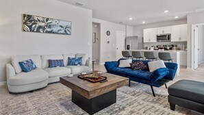 A modern living room with a white sofa, a blue sofa, and a wooden coffee table. There is a kitchen in the background with white cabinets and a microwave.