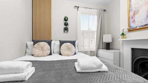 A neatly arranged bedroom with a large bed, a bedside table, a lamp, a fireplace, and a window with curtains.