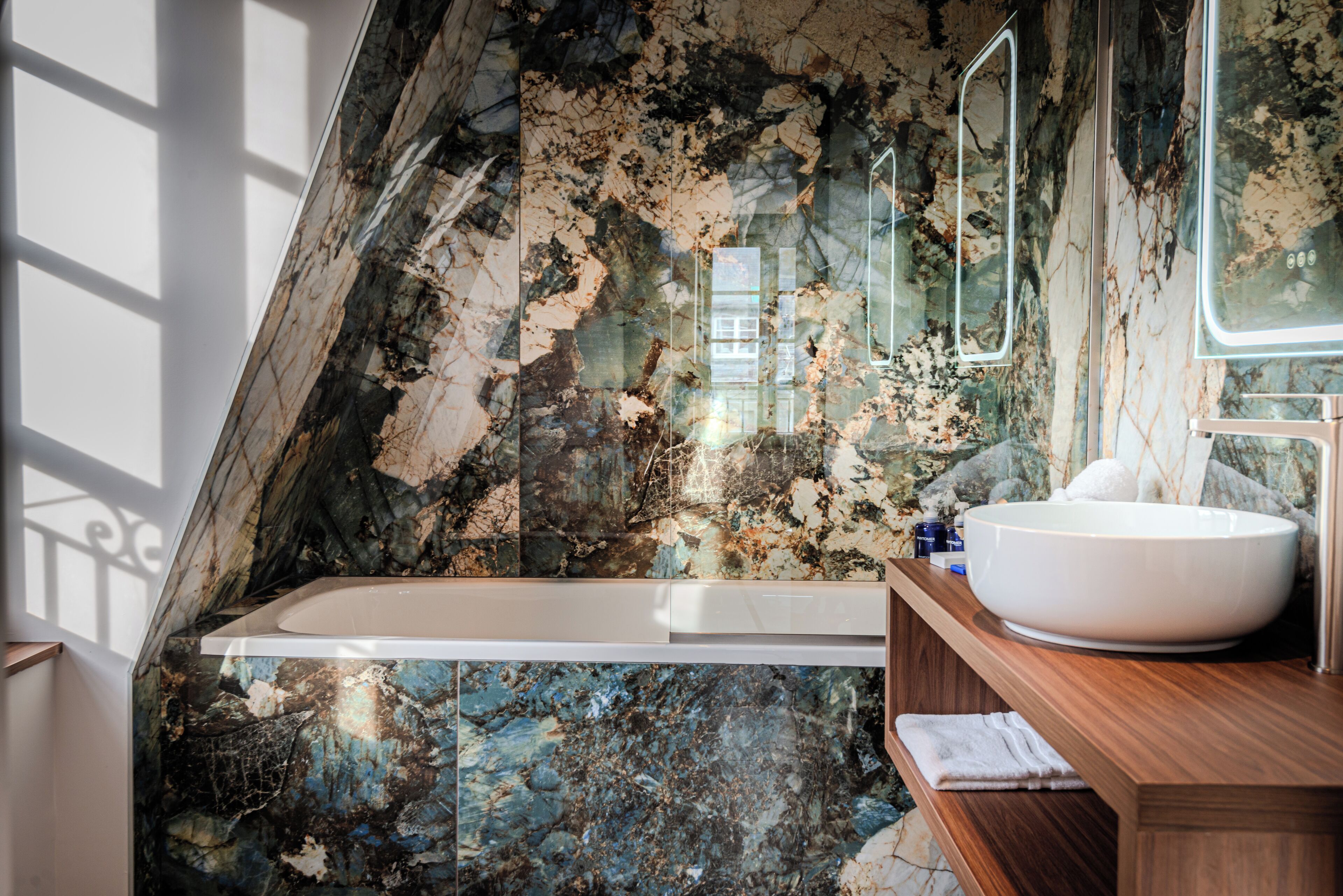 A bathroom with a marble wall, a bathtub, a wooden vanity, and a large mirror.
