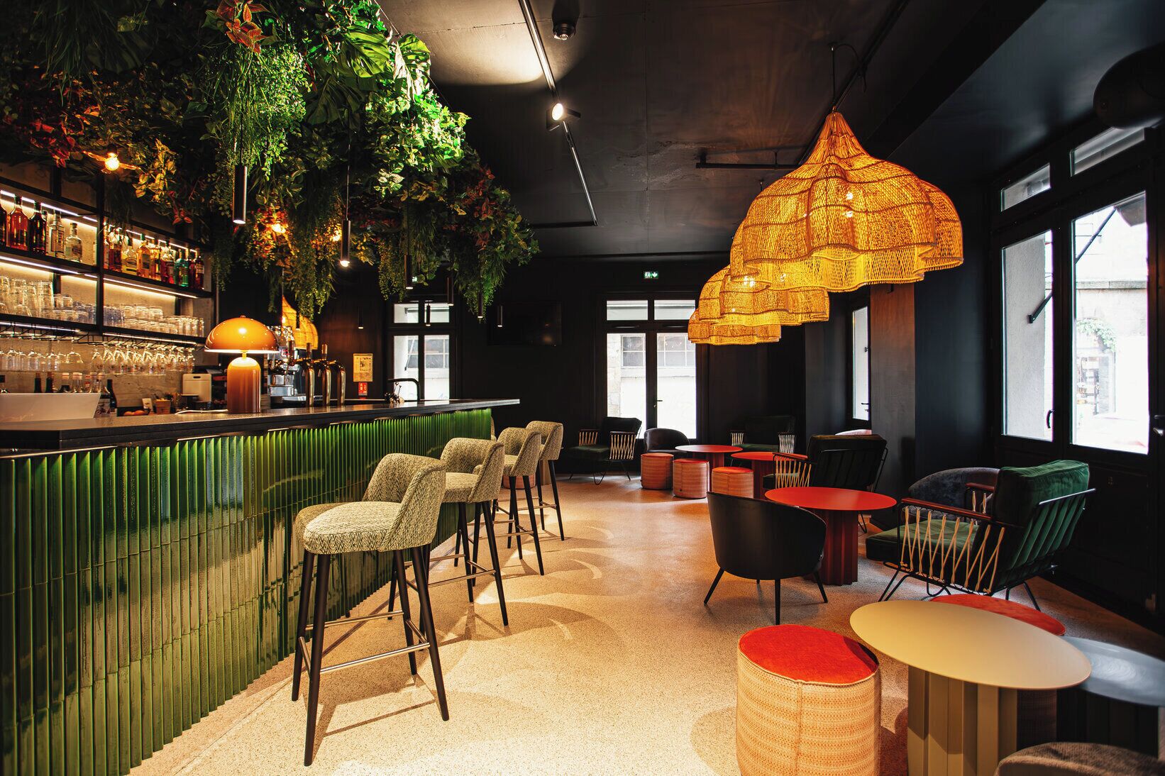 A bar area with a green counter, multiple bar stools, and hanging plants. There are tables and chairs for seating, and a large, textured hanging light fixture above.