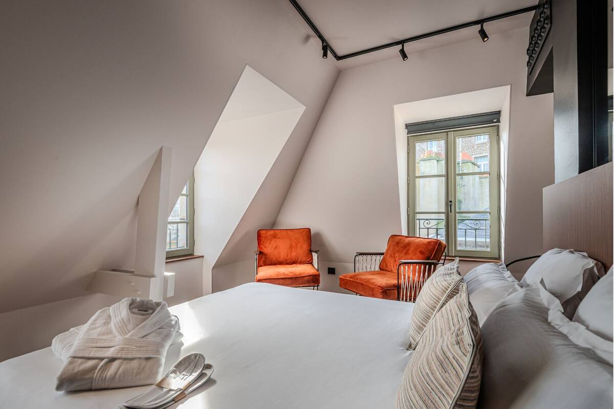 A modern bedroom with a sloped ceiling, a large bed, two orange chairs, and a window with a view of buildings.