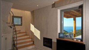Una escalera lleva a un rellano con chimenea, un cuadro enmarcado y un ventanal con vistas al mar.