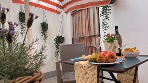 A dining area with a table set for two, a wicker chair, a woven table runner, hanging plants, and a striped umbrella. - CORNABRUGIA: Modern Open Space with Private Patio - AC & 2' to Beach (Vernazza)