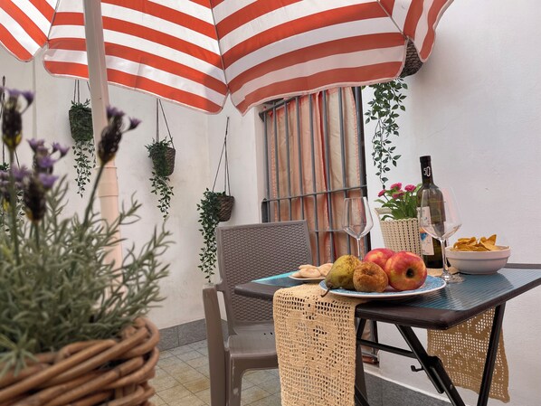 A dining area with a table set for two, a wicker chair, a woven table runner, hanging plants, and a striped umbrella. - CORNABRUGIA: Modern Open Space with Private Patio - AC & 2' to Beach (Vernazza)
