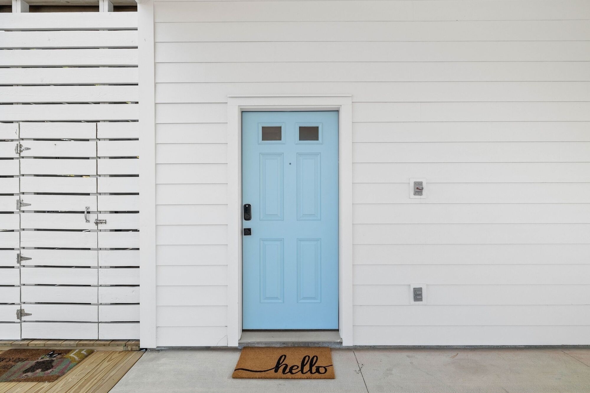 A light blue door with a small window, a doormat with "hello" on it, and a white exterior wall with horizontal siding.