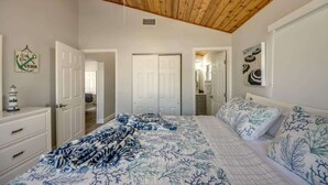Un dormitorio con techo de madera, una cama con colcha floreada azul y blanca, un aparador y una puerta que da a otra habitación.