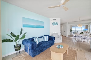 Una sala de estar con un sofá azul, una mesa de centro, una planta grande y un cuadro de una playa.