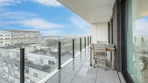 A balcony with a glass railing, a wooden bench, and a view of other buildings.