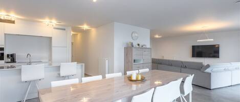 Une cuisine moderne avec des meubles blancs, un coin repas comprenant une table en bois et des chaises blanches, un canapé gris et un téléviseur à écran plat fixé au mur.