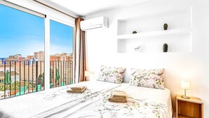 Una camera d'albergo con un ampio balcone, un letto con lenzuola a motivi floreali, un condizionatore d'aria a parete e un comodino con una lampada.